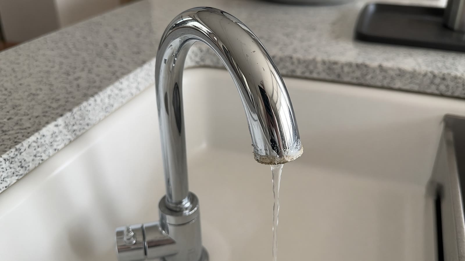 A chrome kitchen faucet with mineral scale on the spout and a thin stream of water.